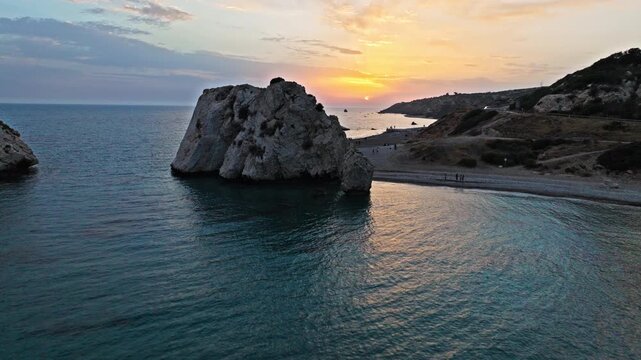 Aerial sunset view of Petra tou Romiou at golden sunset in Cyprus. The iconic rock formation rises from the calm sea, creating a dramatic Mediterranean landmark view.