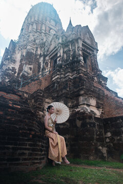 Portrait of Asian teenage girl dressed in traditional Thai outfit and umbrella with cheerful smile at old brick wall castle, holiday travel in Ayutthaya, ancient ruins heritage,  tourist destination.