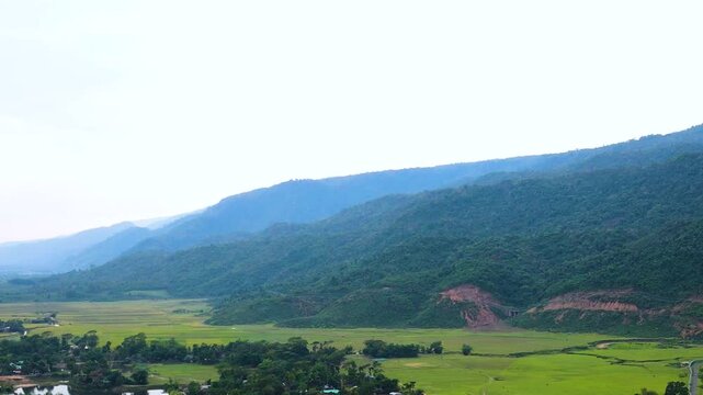 Farmland and village, agricultural fields spread below the lush Meghalaya hills near the Bangladesh India border area, hazy blue sky, daytime, high angle wide aerial drone shot, natural rural location