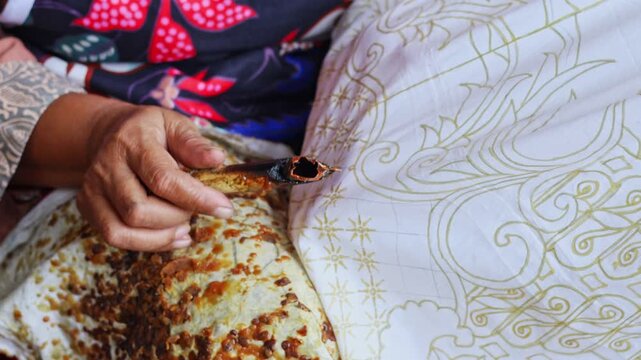 Close-up video of Indonesian craftswoman painting detailed designs on plain cloth during traditional handmade batik workshop