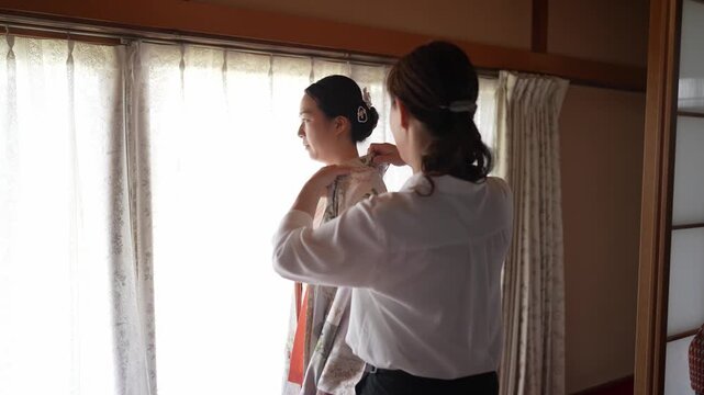 Soft light filters through sheer curtains. Two women prepare for a traditional moment. One carefully adjusts a patterned kimono. Serene focus, quiet concentration. Ideal for cultural, bridal