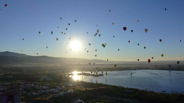 Cinematic drone aerial shot,wide-angle view of dozens of colorful hot air balloons floating in a clear blue sky above a lake at sunrise,mountain backdrop, golden hour light, morning haze over the city