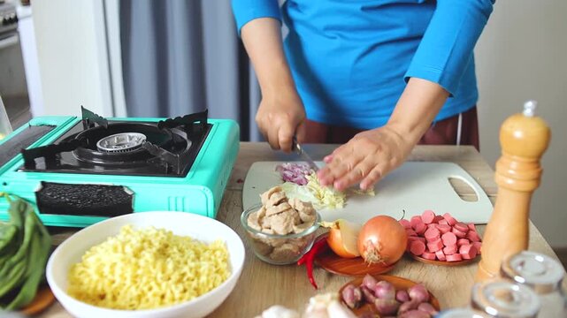 Chef mincing garlic on cutting board next to portable gas stove and cooking ingredients