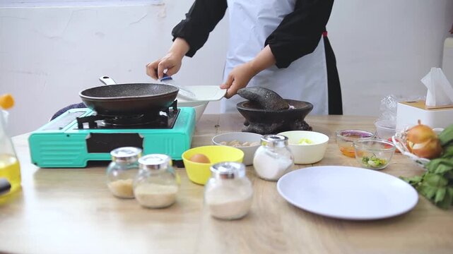 Person wearing apron uses mortar and pestle to grind shallots and garlic on wooden table
