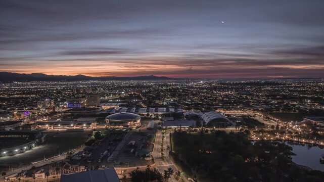 Twilight sky with orange purple gradient, city lights turning on,with curved roof structures and lake visible, illuminated avenues with car traffic, urban sprawl and architectural landmarks.