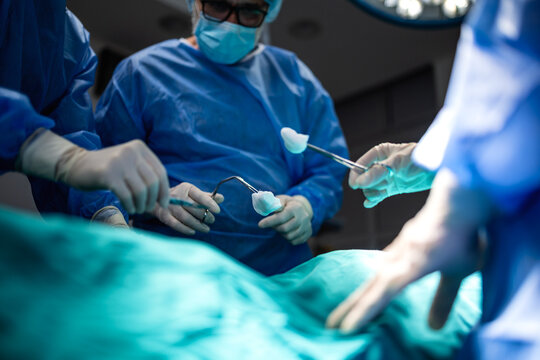 Medical team hands performing surgical operation in a modern hospital room. Professional doctors in blue gowns and sterile gloves using surgical instruments over a patient covered with green sheet