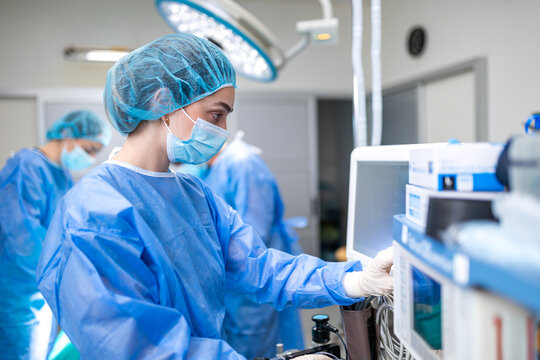 Female doctor in blue surgical scrubs and protective mask operating a modern medical monitor in an operation room. Medical staff working during a surgical procedure under bright surgical lights