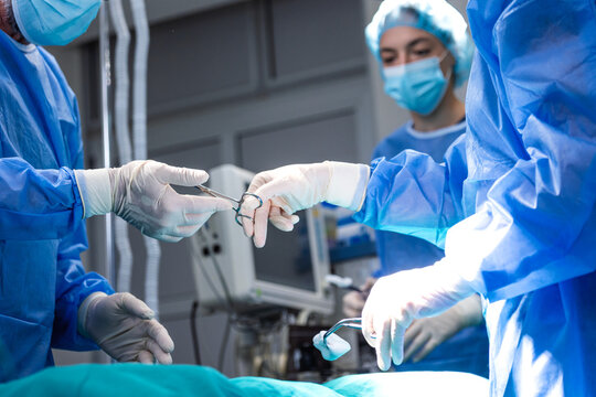 Surgeon passing surgical instrument to assistant during an operation in a hospital operating room. Medical team collaborating in blue scrubs and sterile gloves during a surgical procedure