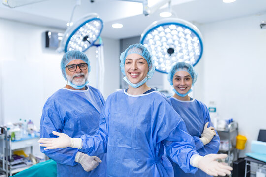 Smiling medical team of surgeons and nurses in blue scrubs and caps posing together inside a bright modern hospital operating room with surgical lights overhead. 