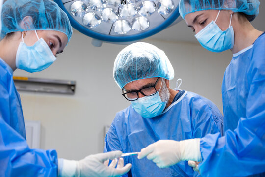 Experienced surgeon passing a surgical tool to an assistant nurse during an operation inside a hospital operating theater. Medical professionals in blue scrubs and sterile gloves working together