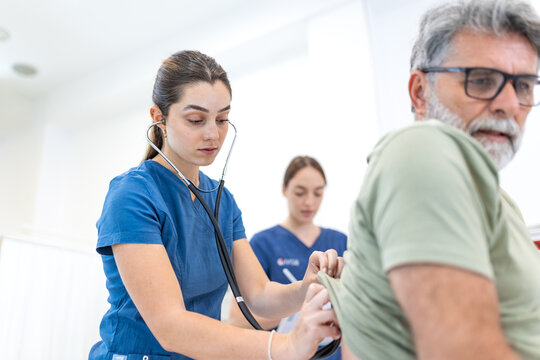Young female doctor using a stethoscope to listen to an elderly male patient's back lungs during a medical checkup inside a bright hospital room, with another doctor in the background.