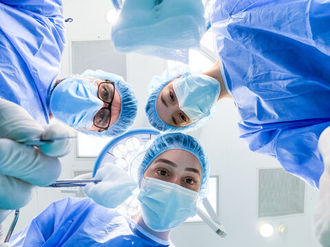 Low angle point of view of a surgical team looking down at the camera wearing blue scrubs, caps and masks in a hospital operating room, ready to perform a medical procedure.