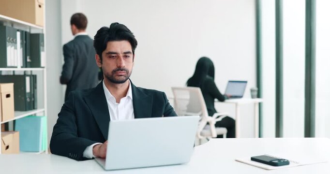 Middle Eastern businessman sits at desk with laptop as colleague walks by. Focused man types on keyboard carefully checking screen during corporate office task