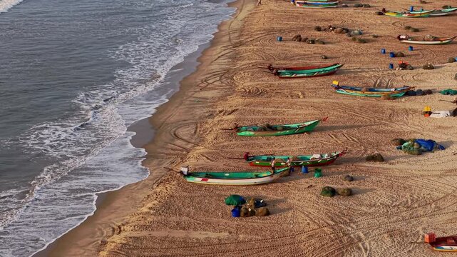 Aerial drone shot of colorful fishing boats resting on the sandy beach as waves wash onto the shoreline along the Bay of Bengal near Chennai in Tamil Nadu, India