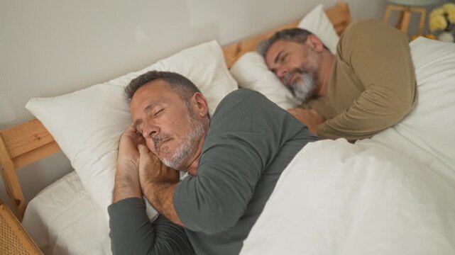 Man touching partner's shoulder and resting a hand on his back on a bed in a bedroom, one partner asleep while the other leans in to kiss; intimacy comfort.
