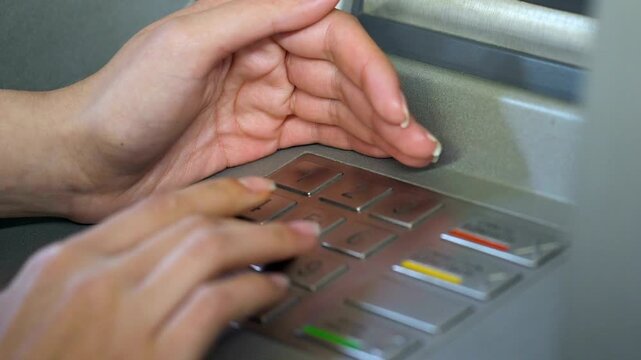 Female entering her pin code and hiding keyboard of automated teller machine with hand for safety and privacy compliance in 4K Ultra HD footage
