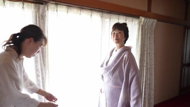 Soft light bathes a traditional Japanese room. Woman smiles, draped in a pale kimono. Another woman adjusts the garment with care. Their calm expressions convey gentle focus