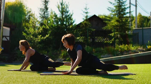 Elderly women practicing gentle Pilates stretches together outdoors on yoga mats, performing slow motion flexibility exercises in a peaceful green garden setting