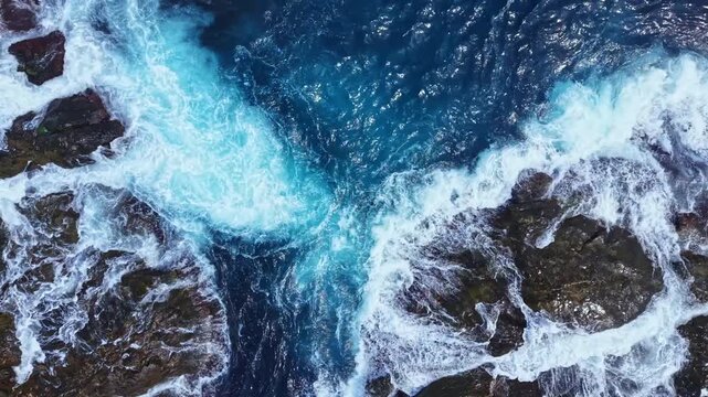 Waves roll and crash against rocky surfaces in a coastal area. The water shows varied shades of blue and white foam as it moves. The scene takes place during bright daylight.