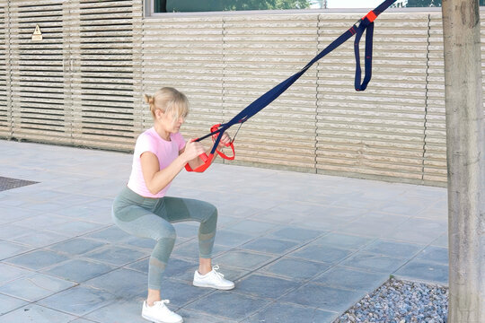 Young woman performing suspension training squat outdoors using resistance straps anchored to wooden post beside modern patio, focused posture and controlled balance in daylight