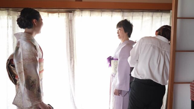 Three women in a Japanese room near a window. One woman wears a patterned kimono, standing still. Another adjusts a pink kimono on a third woman. The room has wooden framing and soft light
