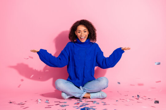 Young mixed race woman in blue sweater sits on pink background smiling with confetti expressing joy fashion lifestyle and celebration moment