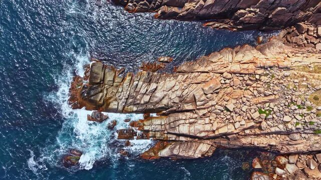 The photo shows a rocky shoreline by the ocean. Waves crash against the rocks creating white foam. The scene captures natural textures of the stones and clear blue water.
