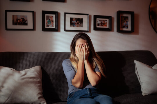 Sad Woman Crying On Couch Covering Face With Her Hands. Emotional depressed female sits alone weeping silently while hiding behind palms indoors
