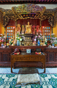 Golden monk statue on an ornate wooden altar at Phap Bao Pagoda or Chua Phap Bao Buddhist temple in Hoi An, Vietnam, featuring ritual offerings, flowers, and a prayer cushion for devotees.