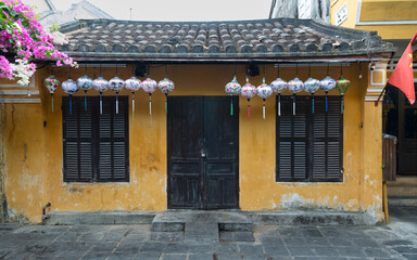 Fototapeta premium Traditional yellow house with dark wooden shutters and colorful silk lanterns in the Ancient Town of Hoi An, Vietnam, adorned with blooming pink bougainvillea and a small national flag