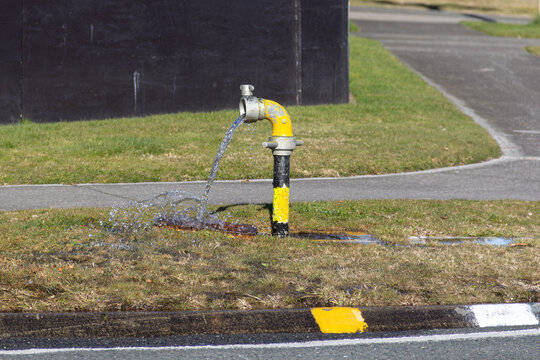 Fire hydrant standpipe, yellow painted, "uncontrolled" style. Water flow is routine maintenance to ensure water quality and system functionality. In Taupō, New Zealand.