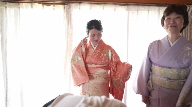 Soft light glows through sheer curtains. Two women dress in kimonos, one adjusting. Third smiles warmly beside them. Gentle moments of traditional Japanese culture