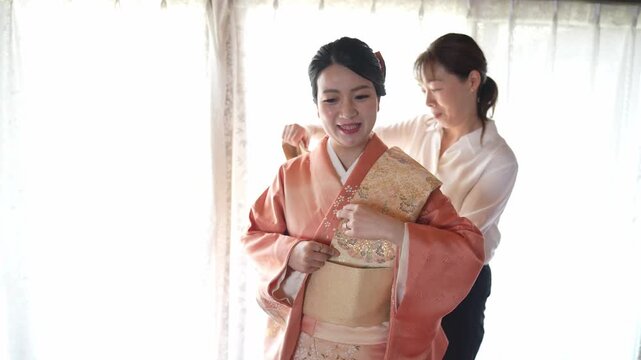 Soft light filters through sheer curtains behind two women. One woman adjusts a peach kimono with gold embroidery. Another helps her with gentle care and precision. The moment radiates calm