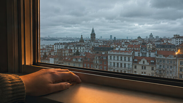 Close-up of a person hand resting on a windowsill, looking out at a grey overcast autumn sky over an unfamiliar foreign city skyline. Adjustment and distance from home concept.