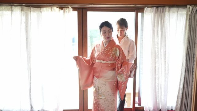 Soft light fills a traditional Japanese room. Woman adjusts a vibrant kimono for another. The second woman stands quietly, dressed in pink. Their focused actions suggest care and preparation