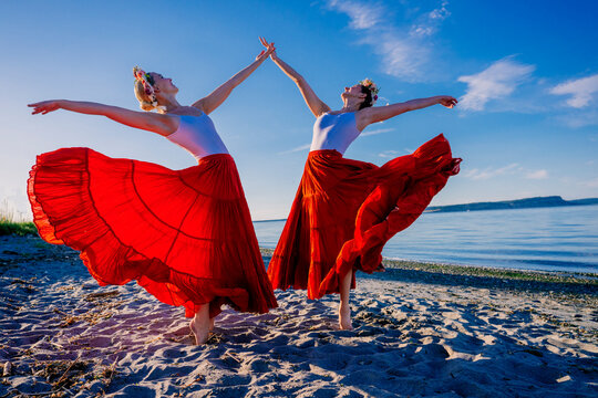 Two white women dancing gracefully on a beach with flowing red skirts