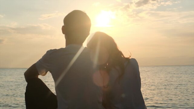 Back view, teenage couple sits side by side on the seashore watching the sunset. Girl rests her head on the boys shoulder. First love, teenage romance, emotional connection and peaceful moments.