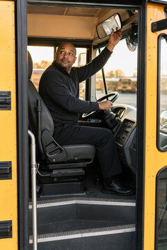 School bus driver in uniform adjusting rearview mirror preparing for commute. Focus on school bus driver in cabin, emphasizing responsibility and safety while transporting students.