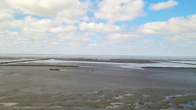Coastal landscape with sandy shore and tidal flats under a bright blue sky filled with fluffy clouds, showcasing the natural beauty of the shoreline