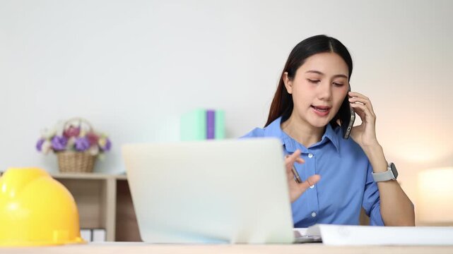 Professional asian engineer working on a laptop computer and talking on the smartphone with a client in her office, multitasking on a construction project with a smile