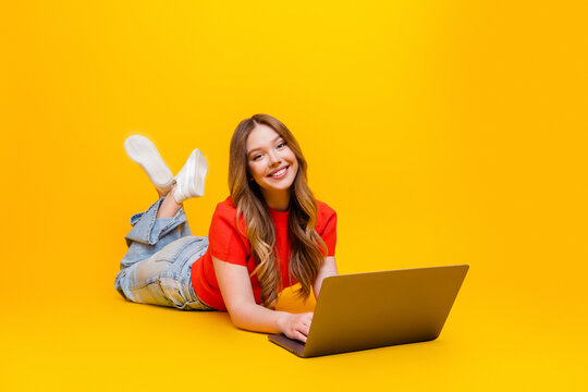 Smiling young woman lying on floor with laptop on bright yellow background, casual remote work and study concept