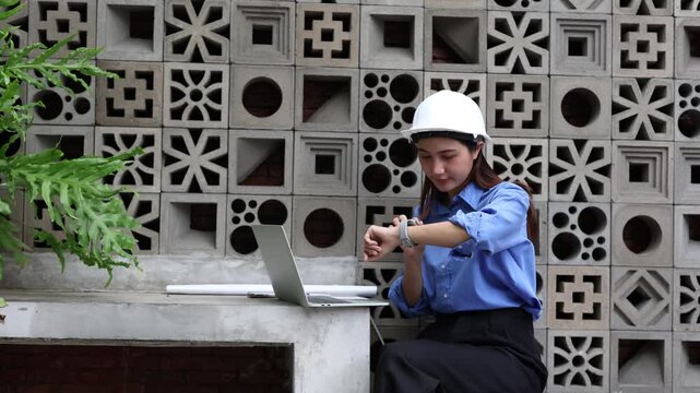 Female civil engineer with a hard hat working on a laptop computer at a construction site, checking her smartwatch while managing a building project with a blueprint