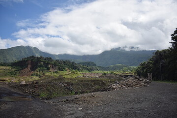 Dirt road winds through rugged terrain. Mist drapes distant, green mountain peaks. Clouds drift softly above the hills. Vast land reveals signs of recent clearing © Fajar