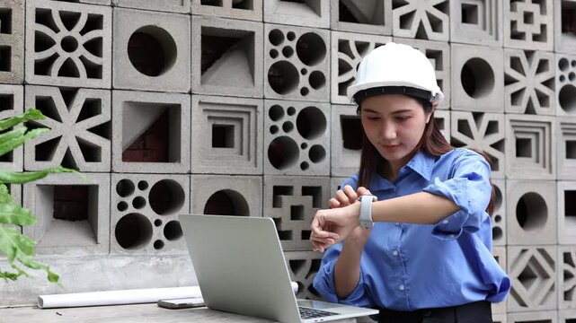 Asian female architect wearing a hard hat working on a laptop at a construction site, checking her smartwatch for notifications and then continuing her design work on the computer