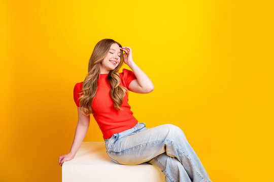 Young woman smiling and relaxed sitting on a white cube against a vibrant yellow background wearing a red top and light jeans