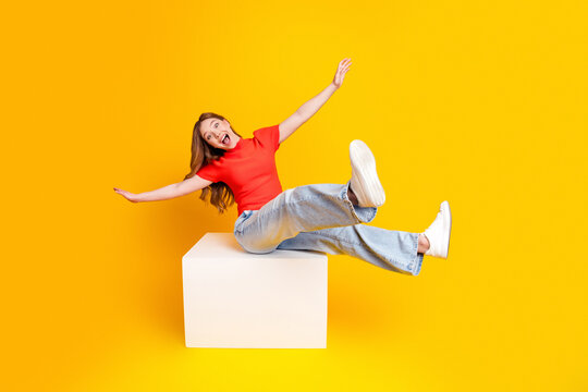 Happy young woman in red top and jeans playfully jumping on white block against bright yellow background, joyful energetic expression and casual fashion studio portrait