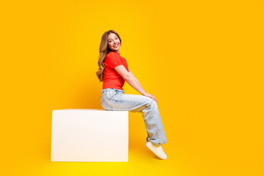 Smiling young woman sitting on a white block against a bright yellow background in casual red top and jeans, cheerful studio portrait conveying happiness, youth and modern lifestyle