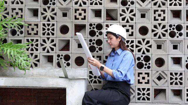 Professional asian female civil engineer with hard hat using laptop and checking blueprint on a construction site, sitting against a concrete block wall background