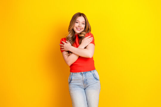 Smiling young woman hugging herself in red top against bright yellow background expressing self love, confidence and happiness