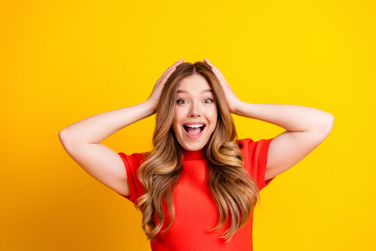 Surprised happy young woman with hands on head wearing red top against bright yellow background expressing excitement and joy